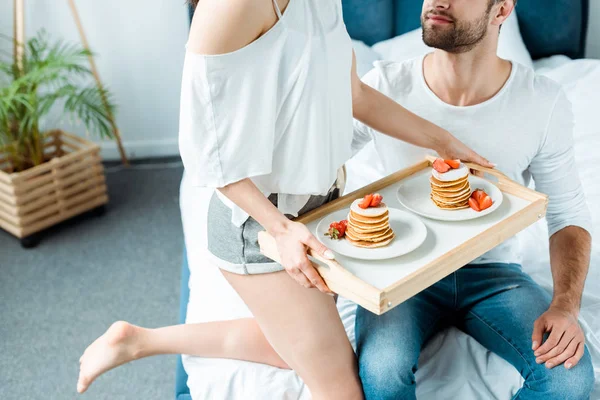 Abgeschnittene Ansicht der Frau mit Holztablett mit leckeren Pfannkuchen und Erdbeeren auf Tellern in der Nähe des Mannes — Stockfoto