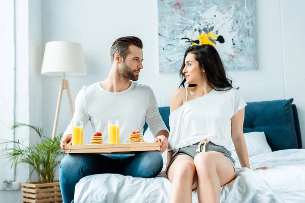 Namorado segurando bandeja de madeira com panquecas saborosas e suco de laranja enquanto olha para a namorada na cama — Fotografia de Stock