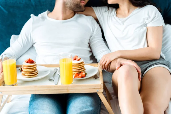 Cropped view of boyfriend holding wooden tray with tasty pancakes and orange juice while touching girlfriend in bed — Stock Photo