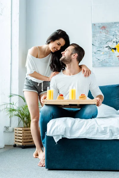 Sorrindo mulher olhando para o homem com suco de laranja e panquecas na bandeja no quarto — Fotografia de Stock