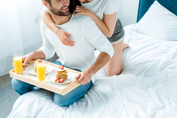 Cropped view of woman hugging man with orange juice and pancakes on tray in bed — Stock Photo