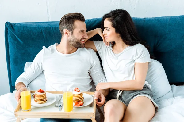 Smiling happy couple having orange juice and pancakes for breakfast in bed — Stock Photo