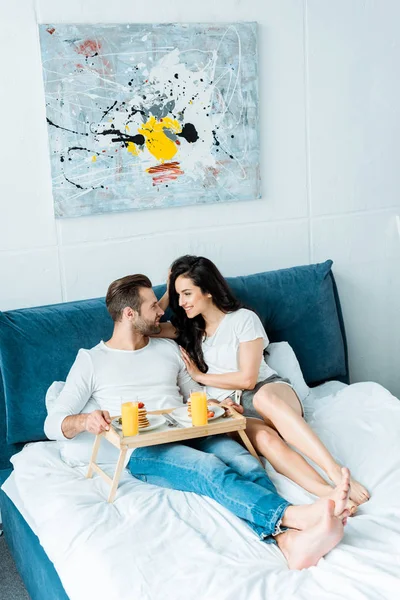 Lächelndes glückliches Paar mit Orangensaft und Pfannkuchen zum Frühstück im Schlafzimmer — Stockfoto