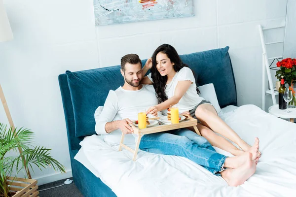 Glückliches Paar mit Orangensaft und Pfannkuchen zum Frühstück im Schlafzimmer — Stockfoto