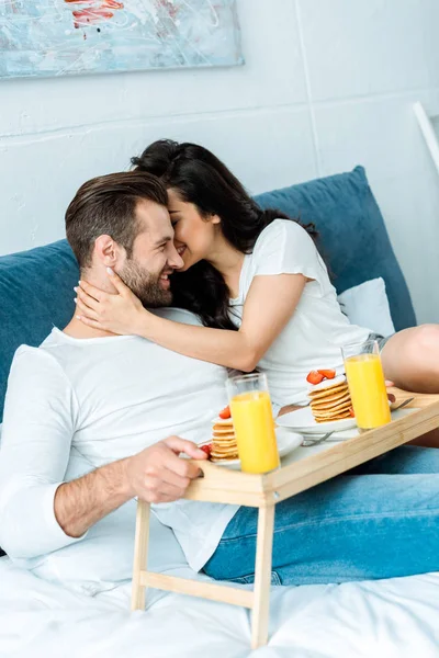 Woman gently hugging man with breakfast on wooden tray in bed — Stock Photo