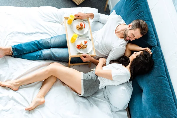 Vista dall'alto della coppia felice a piedi nudi sdraiata a letto con frittelle e succo d'arancia su vassoio di legno — Foto stock