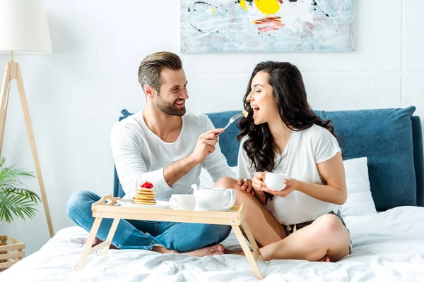 Happy man feeding woman with cup in bed at morning — Stock Photo