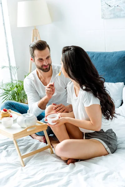 Homem alegre alimentando mulher com copo na cama de manhã — Fotografia de Stock