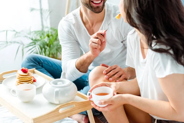 Vista recortada de hombre alegre alimentación mujer con taza de té en la cama por la mañana - foto de stock