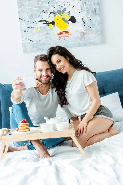 Casal feliz tomando selfie no smartphone perto de bandeja de madeira com café da manhã na cama — Fotografia de Stock