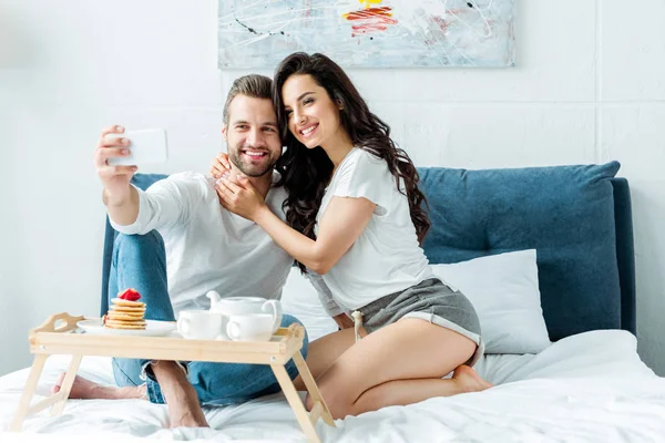 Feliz pareja abrazando y tomando selfie en teléfono inteligente cerca de bandeja de madera con desayuno en la cama - foto de stock