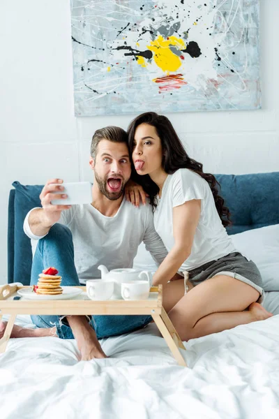 Couple heureux grimacant tout en prenant selfie sur smartphone près plateau en bois avec petit déjeuner au lit — Photo de stock