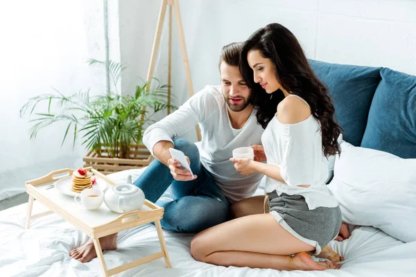 Happy couple having breakfast in bed at morning and using smartphone — Stock Photo
