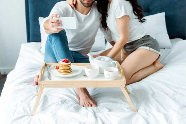 Vista recortada de la pareja tomando selfie cerca de la bandeja con panqueques y té en la cama - foto de stock