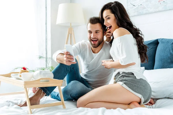 Casal animado tomando selfie perto da bandeja com panquecas e chá na cama — Fotografia de Stock