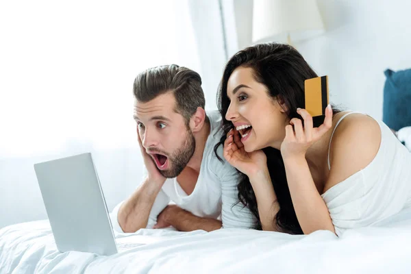 Homme choqué couché dans le lit près d'un ordinateur portable tandis que la femme tenant la carte de crédit — Photo de stock