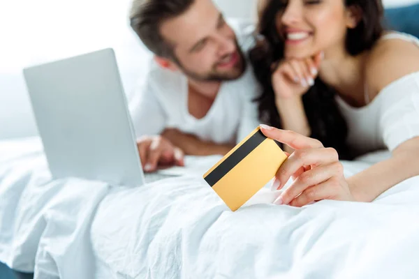 Selective focus of happy woman holding credit card near man with laptop, panoramic shot — Stock Photo