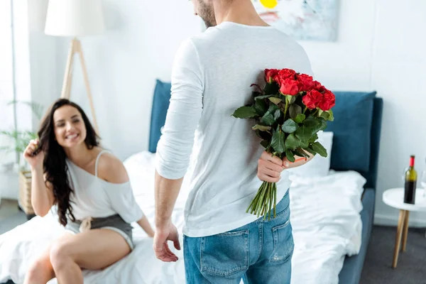 Selektiver Fokus der glücklichen Freundin, die ihren Freund mit roten Rosen auf dem Rücken ansieht — Stockfoto