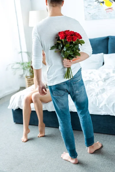 Vista trasera del hombre sosteniendo rosas rojas detrás de nuevo en el dormitorio - foto de stock