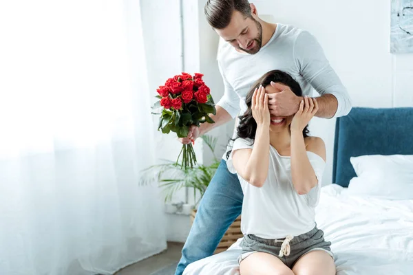 Sonriente hombre con ramo de rosas rojas cerrando los ojos de la mujer para sorprender - foto de stock