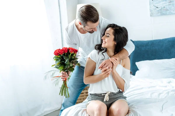 Мужчина с букетом красных роз обнимает улыбающуюся женщину в спальне — стоковое фото