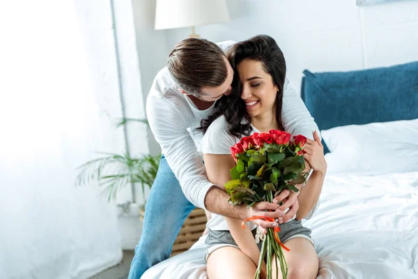 Homem abraçando namorada e buquê de presentes de rosas vermelhas na cama — Fotografia de Stock