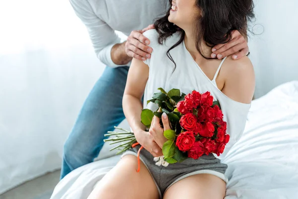 Обрізаний вид чоловіка, який обіймає усміхнену жінку з букетом червоних троянд — стокове фото