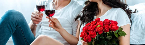 Vue recadrée du couple cliquetant avec des verres de vin rouge et tenant des roses rouges, prise de vue panoramique — Photo de stock