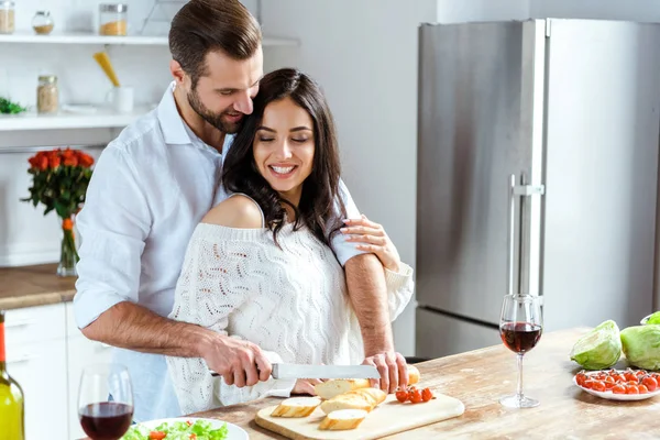 Couple heureux debout ensemble à la cuisine tandis que l'homme coupe le pain sur planche à découper — Photo de stock