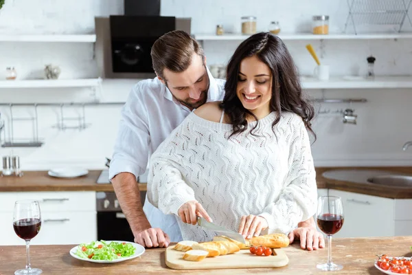 Couple heureux debout ensemble à la cuisine tandis que la femme coupe le pain sur planche à découper — Photo de stock