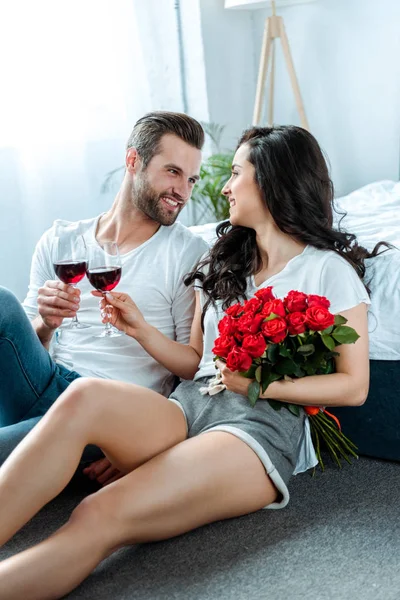 Pareja tintineo con copas de vino tinto y mujer sosteniendo rosas rojas y mirando al hombre en el dormitorio - foto de stock