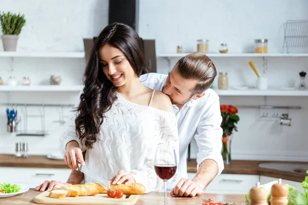 Homem bonito cortando pão na tábua de cortar e beijando ombro namorada — Fotografia de Stock