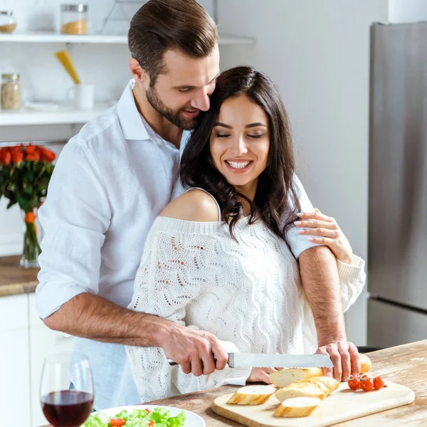 Glückliches Paar, das zusammen in der Küche steht, während Mann Frau umarmt und Brot auf Schneidebrett schneidet — Stockfoto