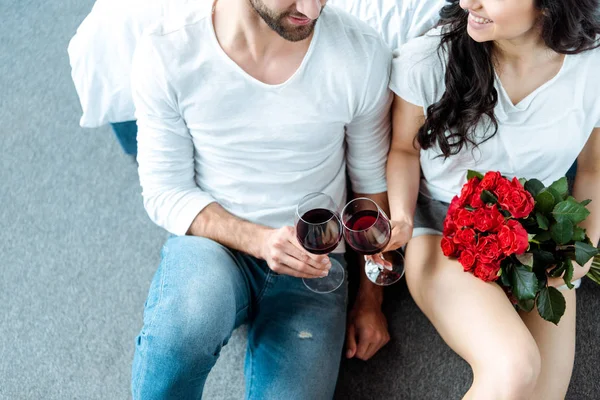 Vista aérea de pareja sonriente tintineando con copas de vino tinto y mujer sosteniendo ramo de rosas rojas - foto de stock