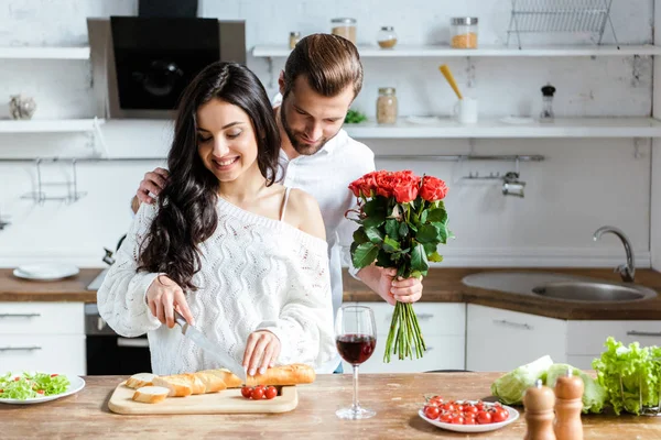 Sonriente pareja de pie juntos en la cocina mientras que la mujer cortar el pan en la tabla de cortar y el hombre sosteniendo rosas rojas - foto de stock