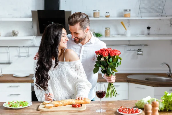 Happy couple cooking dinner while man holding bouquet of roses — Stock Photo