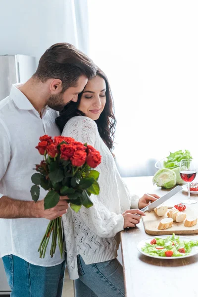 Hombre sosteniendo ramo de rosas mientras abraza a la mujer durante la ensalada de cocina y el corte de pan - foto de stock