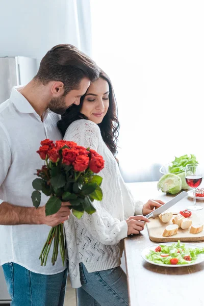 Homem segurando buquê de rosas enquanto abraça a mulher durante a salada de cozinha e corte de pão — Fotografia de Stock