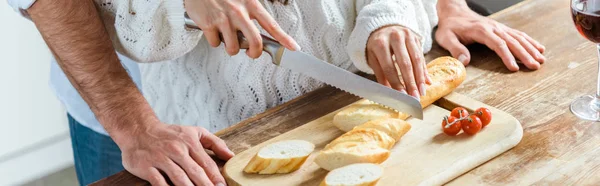 Vista ritagliata dell'uomo in piedi vicino alla donna durante il taglio del pane, colpo panoramico — Foto stock