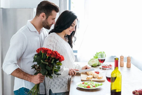 Hombre sosteniendo ramo de rosas mientras abraza a la mujer durante la ensalada de cocina y cortar el pan - foto de stock