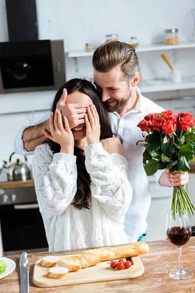 Uomo che tiene mazzo di rose e chiusura occhi donna per fare sorpresa in cucina — Foto stock