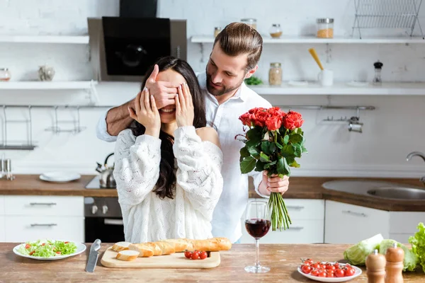 Homem segurando buquê de rosas e fechando os olhos da mulher feliz para fazer surpresa na cozinha — Fotografia de Stock