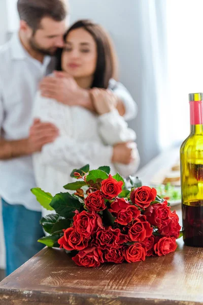 Foco seletivo do homem abraçando a mulher perto do buquê de rosas vermelhas e garrafa de vinho — Fotografia de Stock