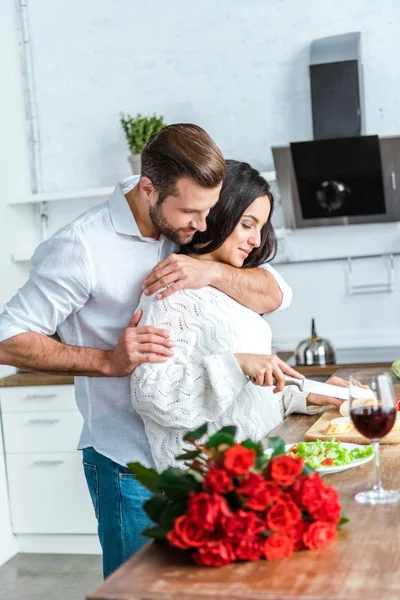 Мужчина обнимает женщину во время приготовления романтического ужина на кухне возле букета красных роз — стоковое фото