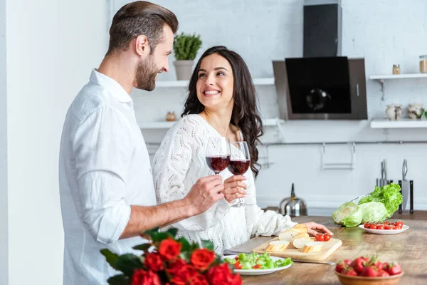Hombre feliz tintineo vasos de vino tinto con la mujer sonriente en la cocina - foto de stock