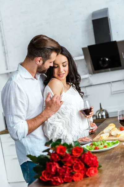 Homem abraçando mulher com copo de vinho tinto na cozinha — Fotografia de Stock