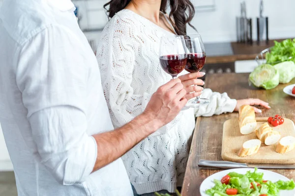 Cropped view of couple clinking glasses of red wine near table with bread and salad — Stock Photo
