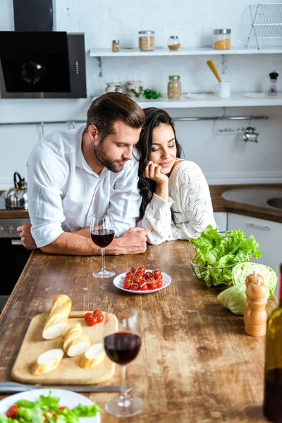 Pareja joven soñadora cerca de mesa de madera con vino tinto, tomates cherry, pan y ensalada - foto de stock