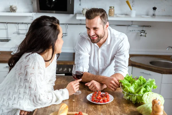 Brünette Frau hält Glas Rotwein neben Holztisch mit Gemüse und schaut glücklichen Mann an — Stockfoto