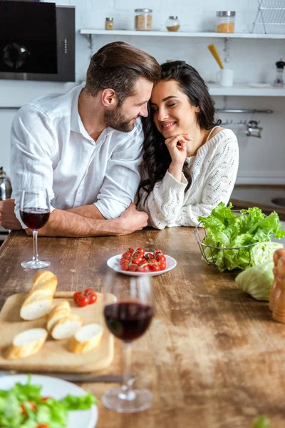 Casal terno sonhador perto de mesa de madeira com vinho tinto, tomate cereja, pão e salada — Fotografia de Stock